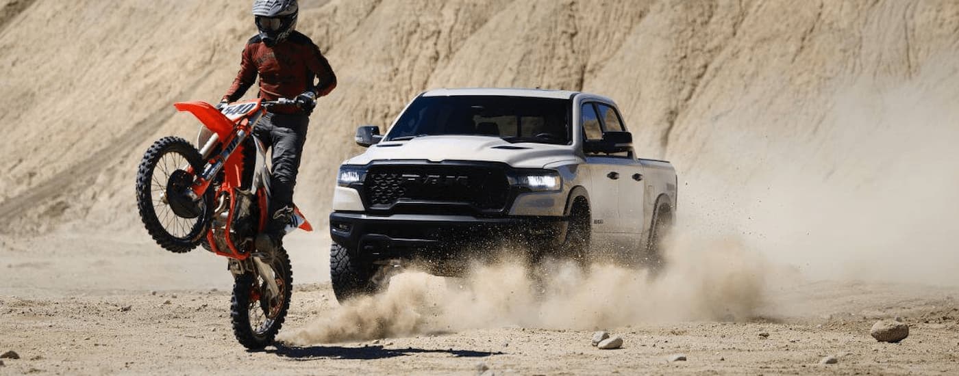 White 2025 Ram 1500 off-roading near a dirt bike in a sand pit